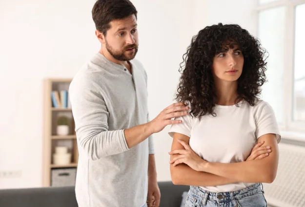 Couple having a disagreement at home as the man tries to talk to his partner, who stands with her arms crossed